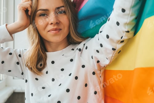 Preview: Young blonde millennial hippie woman with rainbow LGBTQ flag at home balcony. Peace and freedom the