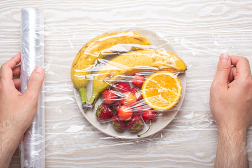 Preview: Male hands wrapping different fruit and berries on white plate in plastic clear film top view on