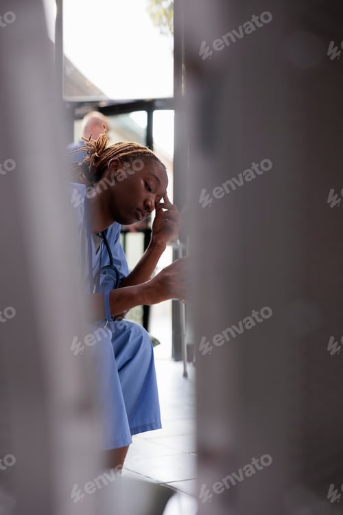 Preview: Tired medical assistant sitting on chair in hospital waiting lobby during checkup visit having a