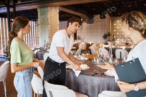 Preview: decorator looking at team lead while arranging festive table setting in modern banquet hall