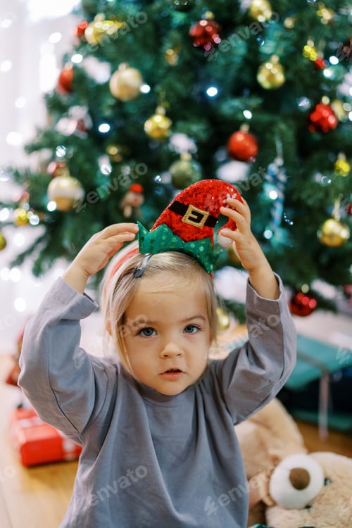 Preview: Little girl tries on a colorful gnome hat on her head while sitting near the Christmas tree