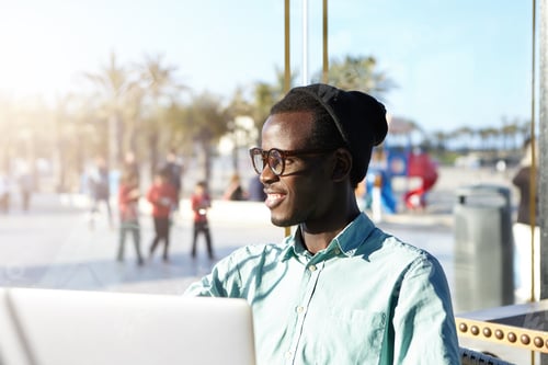 Preview: Stylish African hipster dressed in shirt and black cap sitting in outdoor cafe with laptop checking