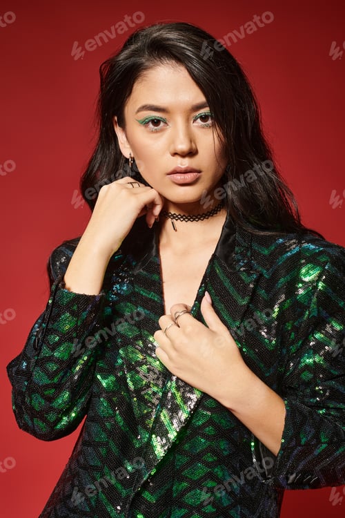 Preview: beautiful asian woman in stylish green jacket with sequins looking at camera on red backdrop