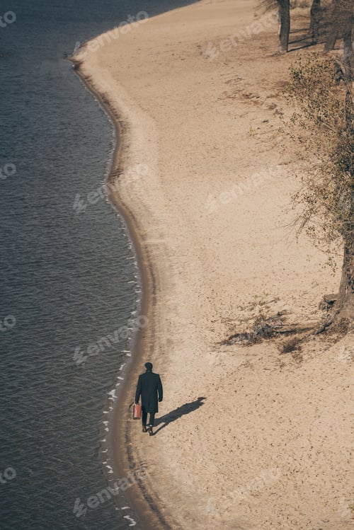Preview: high angle view of man in coat with suitcase walking by sandy beach in autumn
