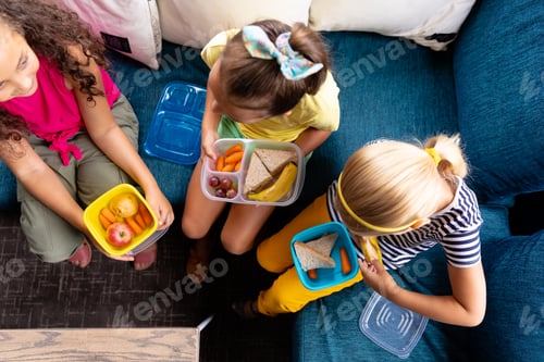 Preview: High angle view of multiracial elementary schoolgirls with lunch boxes sitting on couch during break