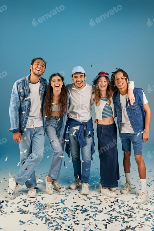 Preview: fashionable multiethnic friends in trendy and denim wear laughing near festive confetti on blue