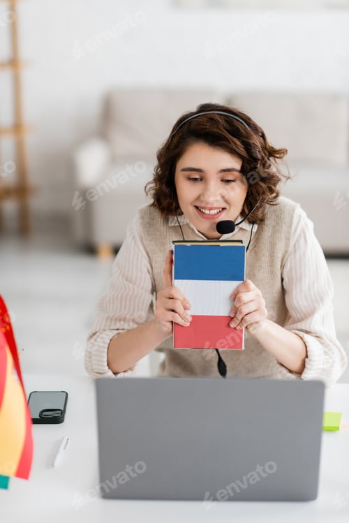 Preview: joyful teacher in headset showing textbook of French language during online lesson on laptop at home