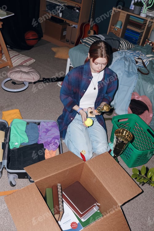 Preview: Caucasian Teenager Girl Packing Trophies and Clothes in Messy Bedroom