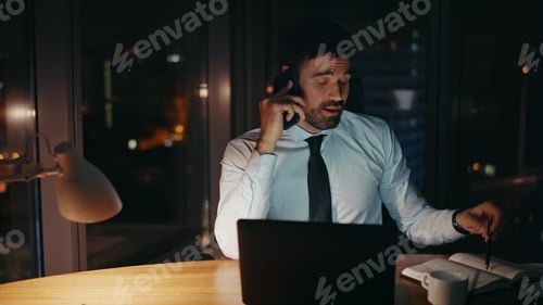 Preview: Overwhelmed employee talking smartphone making notes at office desk late evening