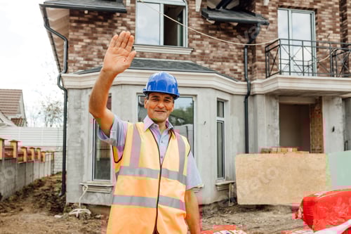 Preview: Attractive cheerful friendly worker in vest and hardhat, waving and saying hello looking at camera.