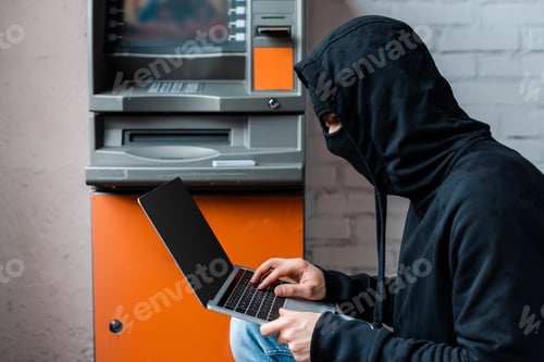 Preview: Side view of hacker in mask using laptop with blank screen near atm