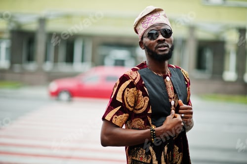 Preview: African stylish and handsome man in traditional outfit and cap standing outdoor.