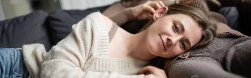 Preview: Smiling woman in sweater looking at camera while lying near girlfriend on couch, banner