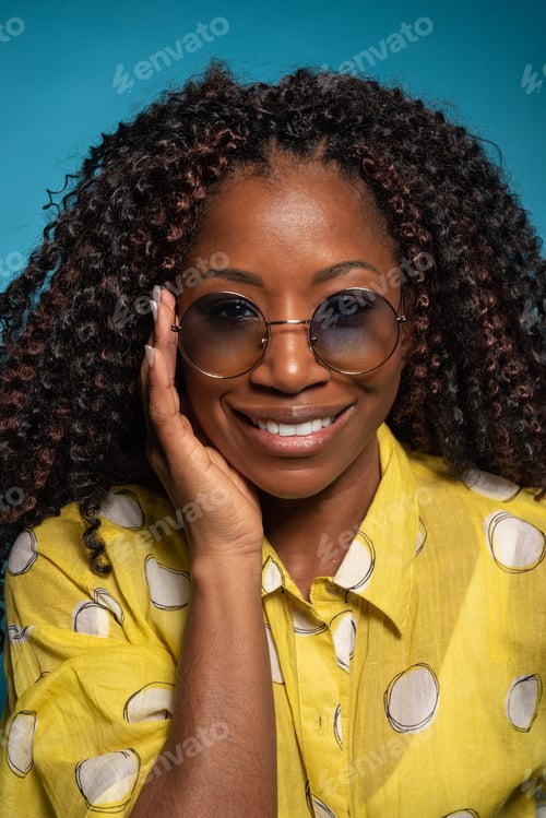 Preview: Close-up of Hispanic woman in sunglasses and yellow shirt smiling at camera on blue background