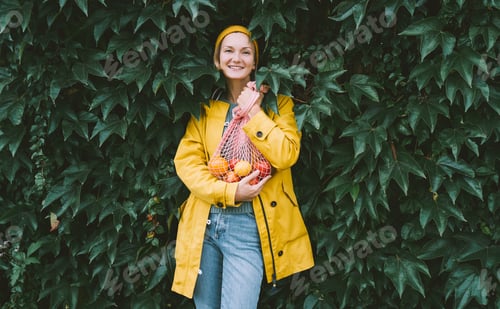 Preview: Young woman wearing yellow raincoat holding bag with organic vegetables against green leaves wall.