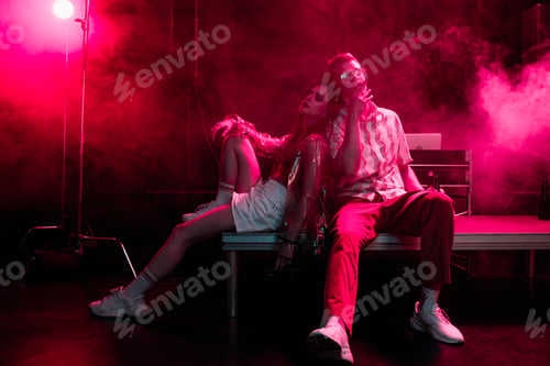 Preview: man and young woman sitting together during rave party in nightclub with pink smoke