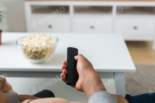 Preview: Hand holds a remote control while a bowl of popcorn sits on a white table in a modern living room