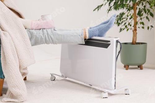 Preview: Young girl in warm knitted socks warms her feet on a radiator. Heating season concept