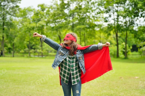 Preview: Happy girl are playing superhero. Kid run across green field in red cloak