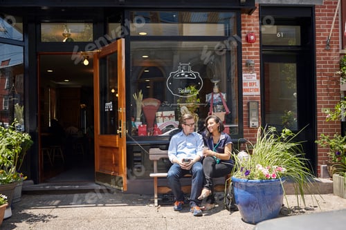 Preview: Couple sitting on bench, outside bakery, holding coffee cups