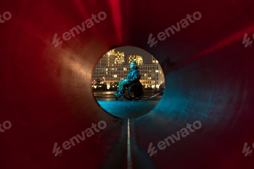 Preview: Young disabled man in rgb color light sits in wheelchair looking at city lights, thinking about life