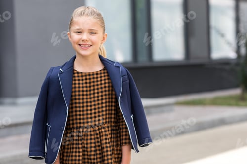Preview: portrait of adorable little blonde girl smiling at camera while standing near school