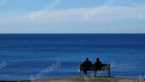 Preview: rear view of two people sitting together on seat at the beach looking out to sea