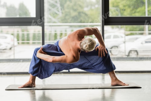 Preview: grey haired man doing wide legged forward fold twist while practicing yoga
