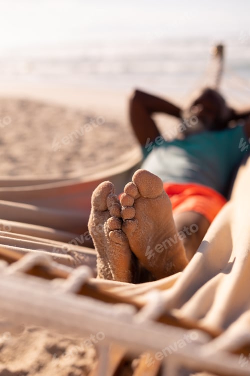 Preview: African american senior man with sand on feet lying on hammock at beach on sunny day