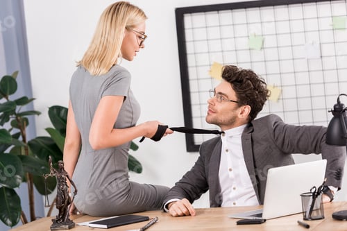 Preview: young businesswoman pulling colleagues tie while sitting on table in office, office romance concept