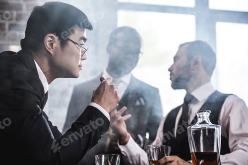 Preview: Multiethnic group of businessmen smoking and drinking whisky indoors, business team meeting