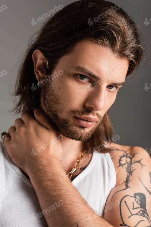 Preview: Portrait of long haired tattooed man touching neck isolated on grey