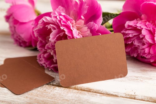 Preview: Brown business card with pink peony flowers on white wooden background. side view, copy space.