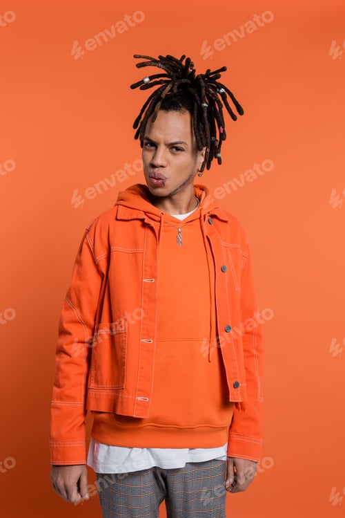 Preview: pierced multiracial man with dreadlocks pouting lips while standing in denim jacket on coral
