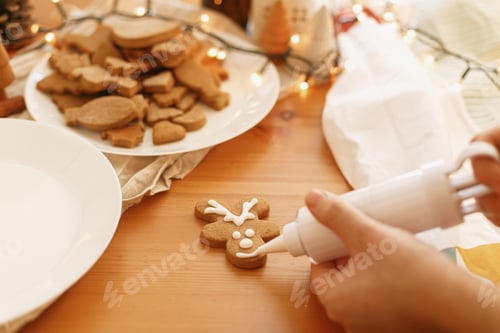 Preview: Decorating gingerbread reindeer cookie with icing on wooden table with lights. Christmas tradition