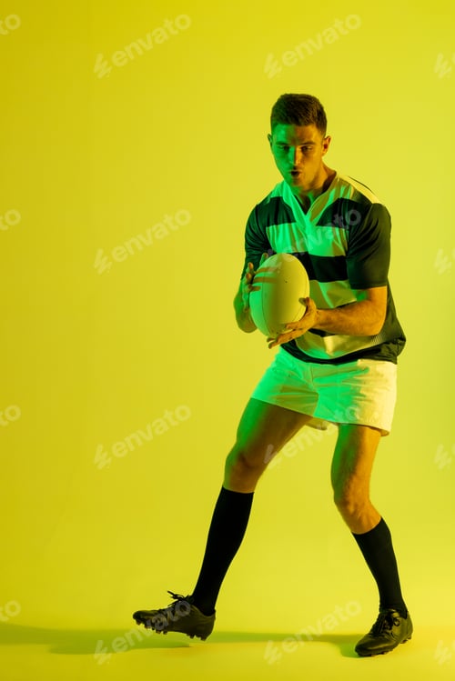 Preview: Caucasian male rugby player catching rugby ball over yellow lighting