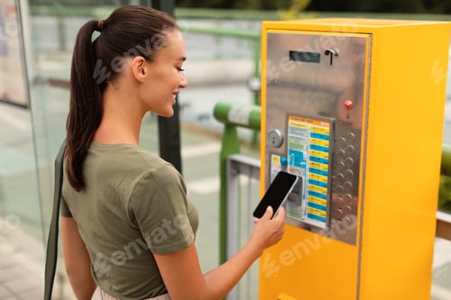 Preview: Young woman standing on bus stop paying ticket with smartphone
