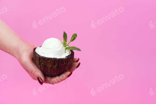 Preview: Vanilla ice cream balls in a fresh coconut half, decorated with mint leaves in your hand on a pink