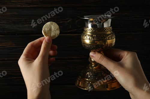 Preview: Liturgical bread and golden cup in hands on wooden background, close up