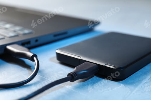 Preview: Hard disk drive with USB cable connected to laptop on blue textured table, closeup