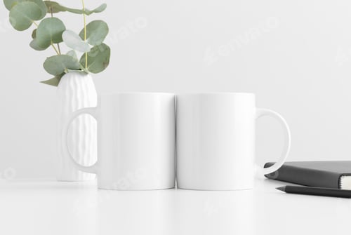 Preview: Two mugs mockup with eucalyptus in a vase and workspace accessories on a white table.