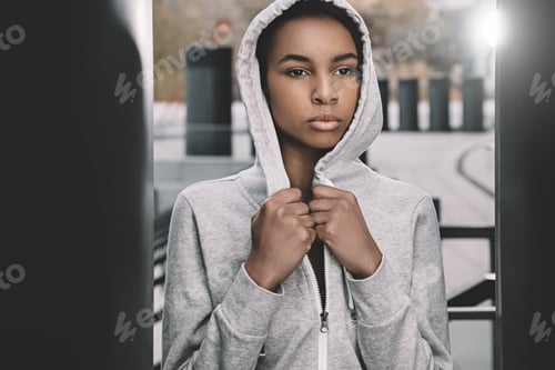 Preview: Attractive young fitness woman in sportswear wearing hood and posing on stadium