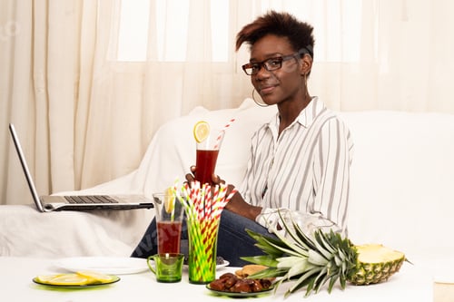 Preview: African woman working on laptop at home near drink and tropical fruits. Remote work