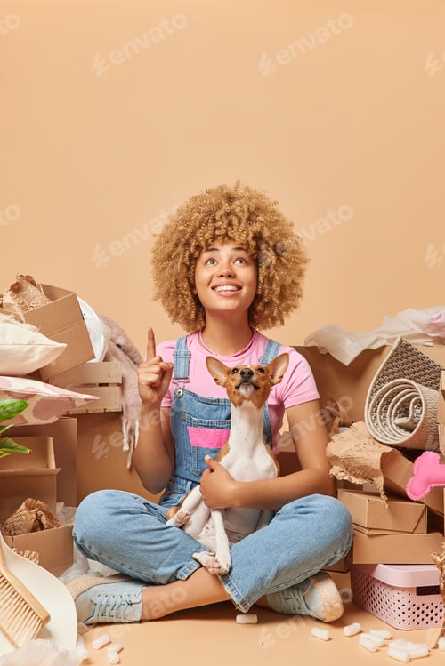 Preview: Happy young woman moves to new apartment with pet sits crossed legs wears t shirt and denim overalls