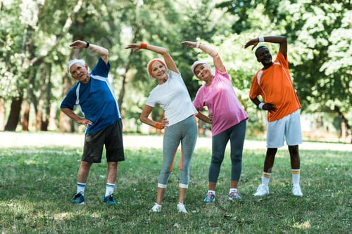Preview: cheerful multicultural retired men and women standing with hands on hips while doing stretching