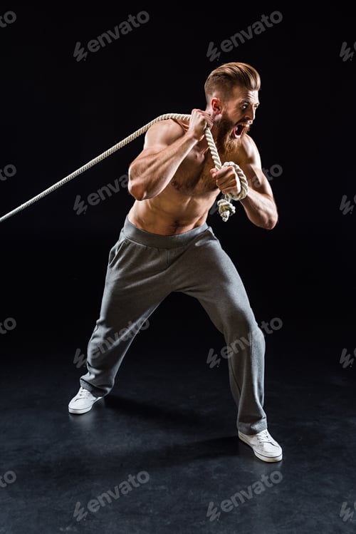 Preview: handsome bearded sportsman yelling and pulling rope isolated on black