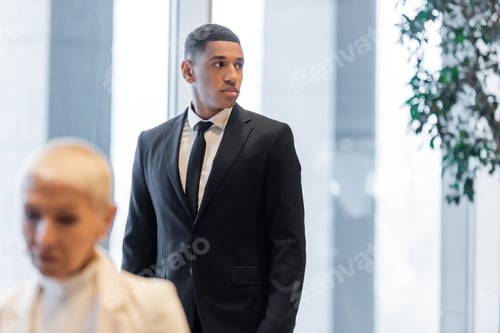 Preview: afrcan american guard in black suit looking away near mature businesswoman on blurred foreground