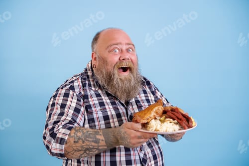 Preview: Happy plump man holds plate of fat food posing on light blue background