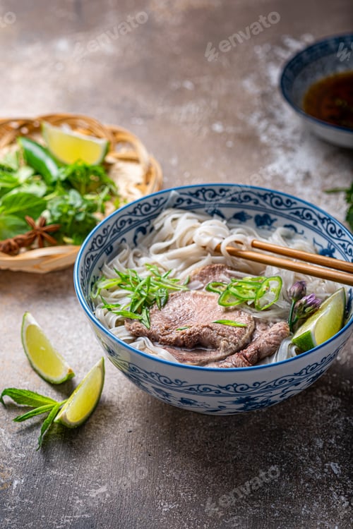 Preview: Close view of pho bo in traditional bowl, garnished with basil, mint, lime, on concrete background