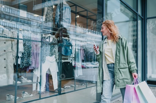 Preview: Woman with shopping bags using smartphone and walking by clothing store
